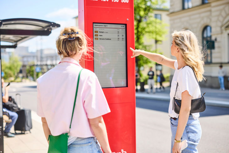 Två kvinnor som tittar på en digital skylt på hållplats Rådhusparken. 