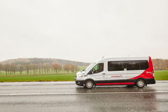 En servicerese-buss kör på en landsväg. Vädret är regnigt och vägen är blöt. I bakgrunden syns åkrar och träd.